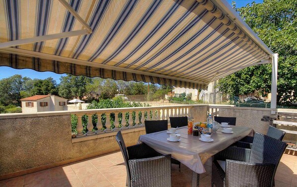 Un patio al aire libre con toldo a rayas, una mesa con vajilla blanca, sillas de mimbre y vistas a edificios y follaje.