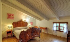 Un dormitorio con una cama de madera, una mesita de noche, una pequeña mesa, una ventana con cortinas y un cuadro en la pared.