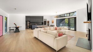 A modern living room with a sectional sofa, a dining table with chairs, a kitchen area, and large windows.