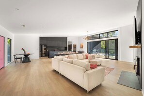 A modern living room with a sectional sofa, a dining table with chairs, a kitchen area, and large windows.
