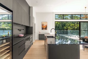 A modern kitchen with a large island, a wine fridge, and a view of a forest through large windows.