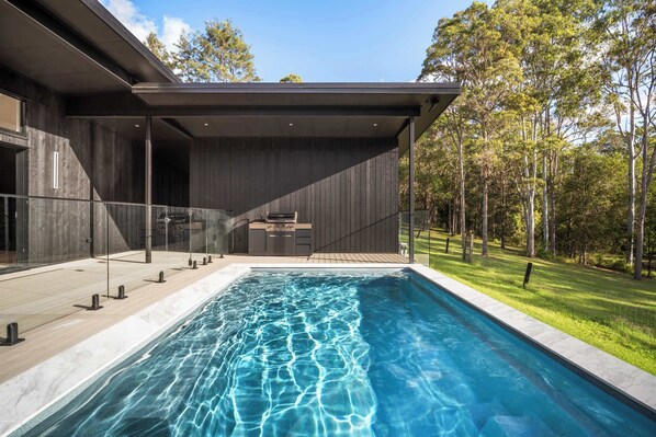 A modern house with a swimming pool, a covered patio, and a view of a forest.