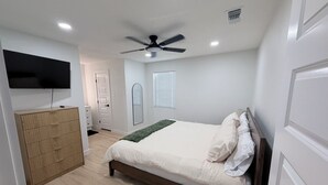Quarto com cama, ventoinha no teto, televisão fixada na parede, cómoda de madeira e porta.
