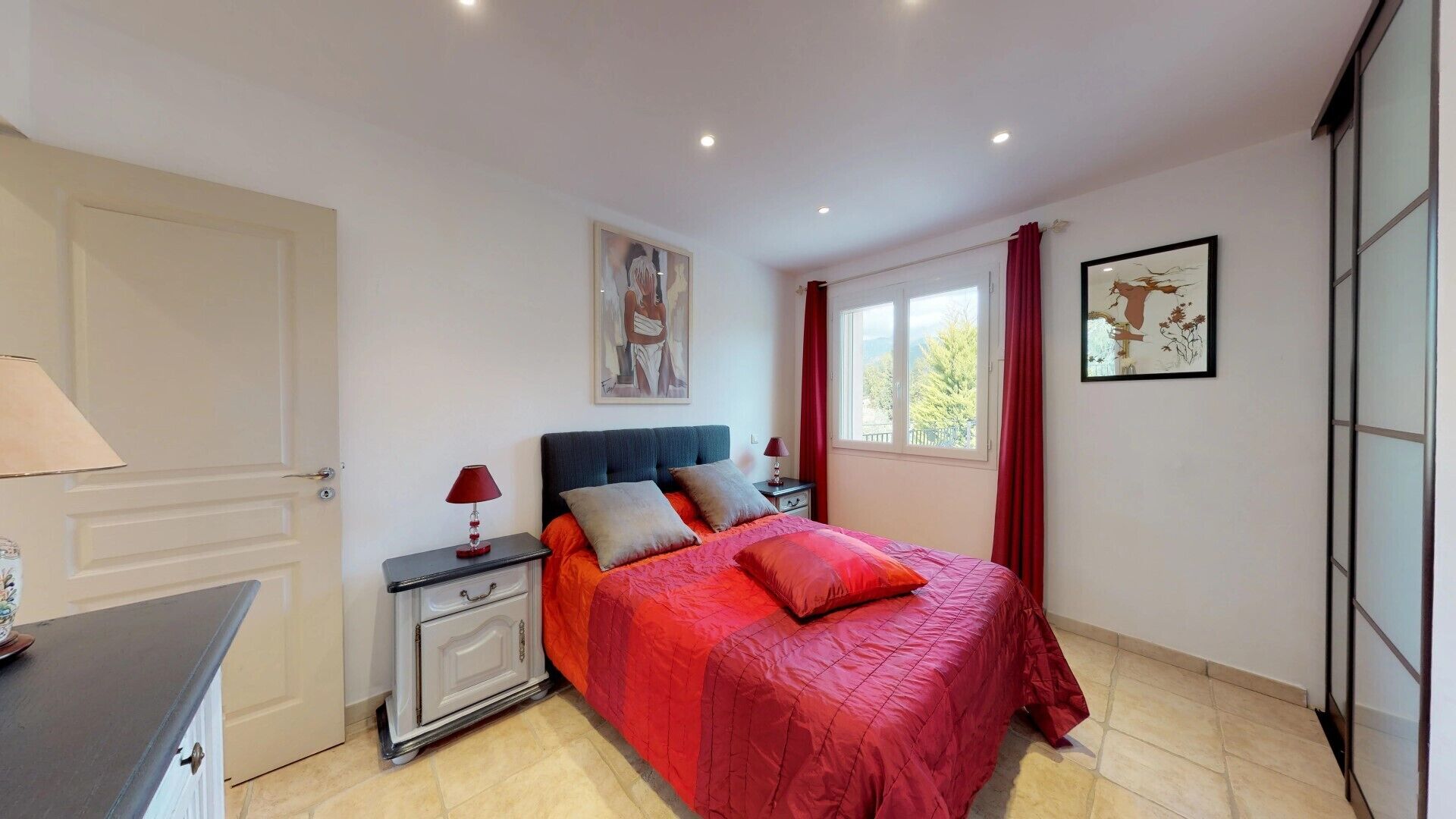 Une chambre à coucher comprenant un lit, des tables de chevet, une commode, une fenêtre avec des rideaux et des œuvres d’art accrochées aux murs.
