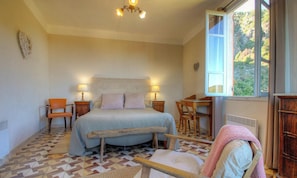 Ein Schlafzimmer mit einem Bett, zwei Stühlen, einem Tisch, einem Heizgerät und einem Fenster mit Blick ins Grüne.