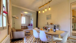 Ein Essbereich mit einem Holztisch, weißen Stühlen und einer Weinflasche. Im Hintergrund befindet sich eine Couch und eine Lampe.