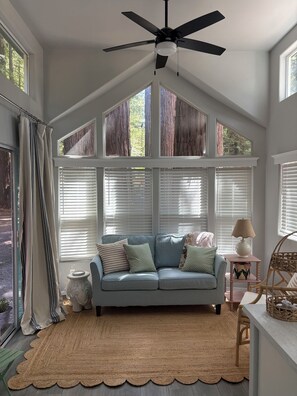 Ein Zimmer mit Deckenventilator, einem hellen Sofa mit Kissen, einem kleinen Tisch mit Lampe und einem Korbstuhl.