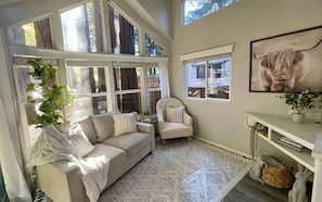 A cozy room with a sofa, armchair, large windows, and a framed picture on the wall.