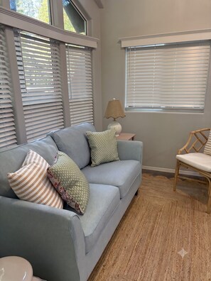 Una habitación con un sofá gris, cojines estampados, una mesita de noche con una lámpara y una silla.