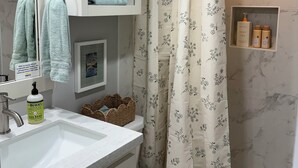 A bathroom with a toilet, sink, mirror, shelves with toiletries, and a shower curtain. - NEW! The Gilded Grove Cottage in Charming Guerneville (Guerneville)