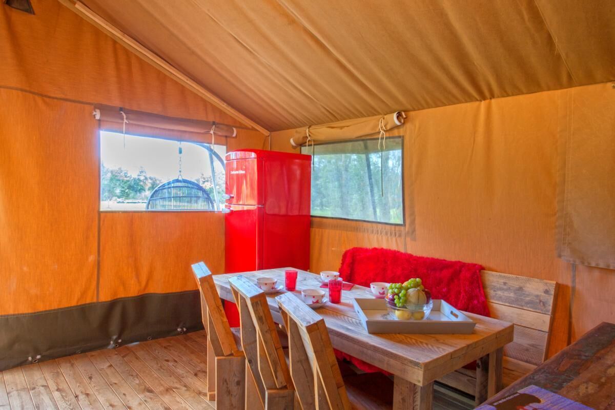 Una habitación tipo tienda de campaña con una mesa de madera, sillas, un sofá rojo y una ventana con vistas a árboles.