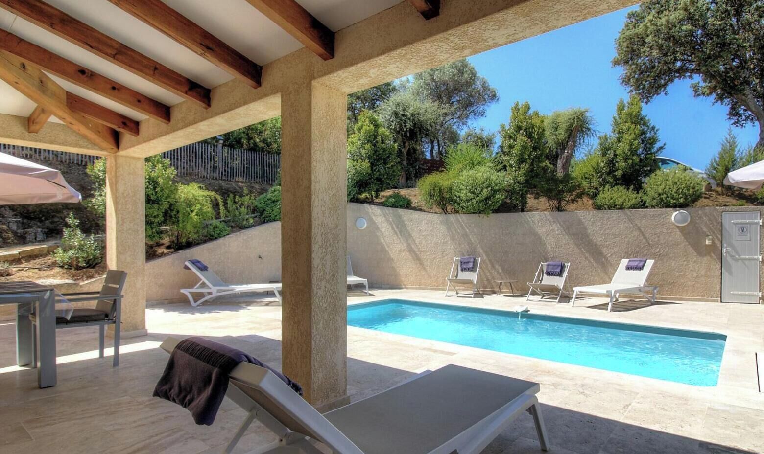 Un espace piscine aménagé avec des chaises longues et une table, entouré d’un mur de pierre et d’arbres.