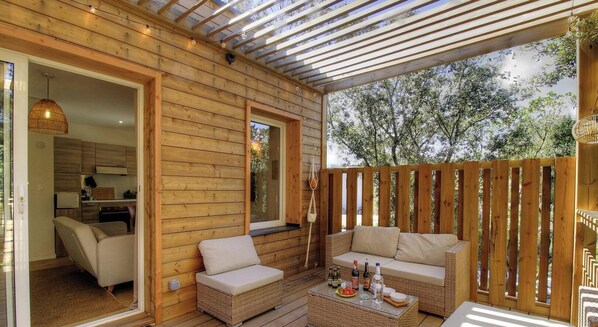 A wooden deck with wicker furniture, a glass table with bottles, and a view of trees. - Pasciale Papanellu - SAINTE LUCIE DE PORTO VECCHIO (SAINTE LUCIE DE PORTO VECCHIO)