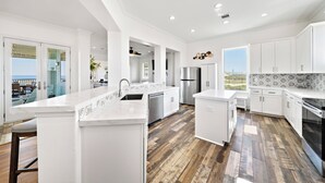 A modern kitchen with white cabinetry, a central island, and a view of the outdoors. - Bay View Beach Home | 6BR | Decks + Views (Galveston)