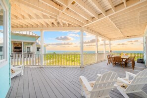 A covered deck with a view of the beach, outdoor furniture, and a dining table. - Bay View Beach Home | 6BR | Decks + Views (Galveston)