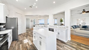 A spacious kitchen with white cabinetry, a central island, and a view to the living area. - Bay View Beach Home | 6BR | Decks + Views (Galveston)
