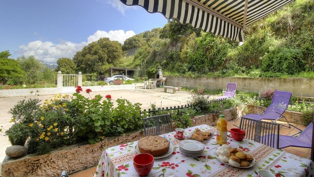 Ein Essbereich im Freien mit Tisch, gedeckt für eine Mahlzeit, umgeben von Grün und Blick auf einen Hügel.