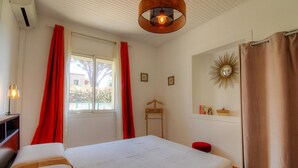 A bedroom with a bed, a window with curtains, a bedside lamp, a wooden chair, a mirror, and a wall-mounted sunburst decoration. - Tarri-Vecchi - PORTO VECCHIO (PORTO VECCHIO)