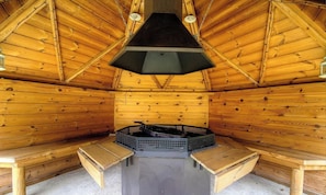 L’intérieur d’un chalet en bois, avec une cheminée centrale, des bancs et un ventilateur de plafond.