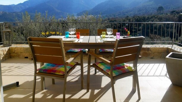 Une table dressée à l’extérieur, avec vue sur la montagne, des chaises en bois ornées de coussins colorés et un parasol jaune.