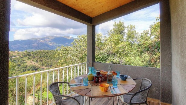 Un área de comedor al aire libre techada con una mesa dispuesta para dos personas, rodeada de sillas, con vistas a un paisaje montañoso.