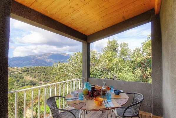 Een overdekte eethoek buiten, met een tafel gedekt voor twee personen, omringd door stoelen en met uitzicht op een bergachtig landschap.