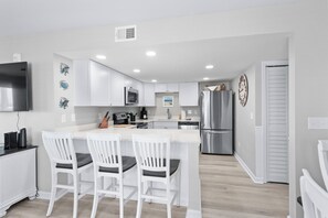 A modern kitchen with white cabinetry, a central island with bar stools, stainless steel appliances, and a flat-screen TV. - Coastal Elegance | Renovated 4BR Oceanfront-Pool & Beach Access (North Myrtle Beach)