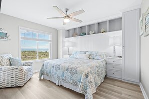 A bedroom with a bed, a nightstand, a chair, a ceiling fan, and a window with a view. - Coastal Elegance | Renovated 4BR Oceanfront-Pool & Beach Access (North Myrtle Beach)
