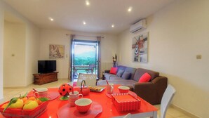 Uma sala de estar com sofá, televisão, mesa de jantar com frutas e copos, e varanda com vista.