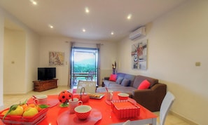 Ein Wohnzimmer mit Sofa, Fernseher, Esstisch mit Obst und Tassen sowie einem Balkon mit Blick.