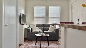 Un espacio habitable compacto con un sofá gris, una pequeña mesa redonda y una cocina con encimera de mármol.