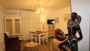 Una habitación con una mesa de comedor de madera, sillas blancas, una mesita redonda con una vela, un sofá, una estatua decorativa, un televisor y una lámpara en el techo.