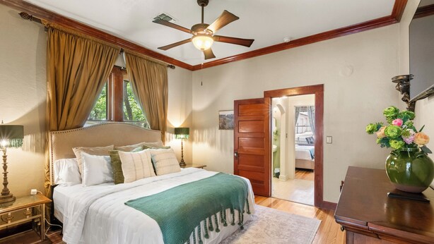 Ein Schlafzimmer mit Bett, Deckenventilator, Nachttisch, Lampe, Fenster mit Vorhängen, Tür und einer Vase mit Blumen.