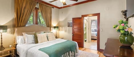 A bedroom with a bed, a ceiling fan, a nightstand, a lamp, a window with curtains, a door, and a vase with flowers.