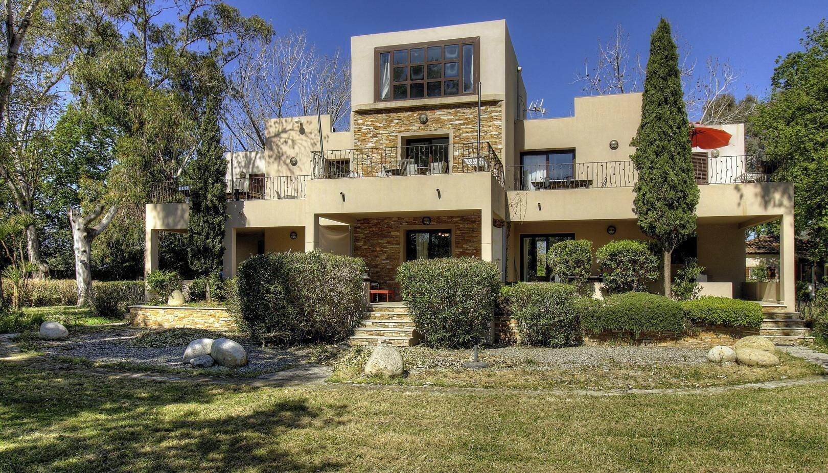 Uma casa moderna de dois andares com fachada de pedra, uma varanda e um jardim bem cuidado com pedras e arbustos.