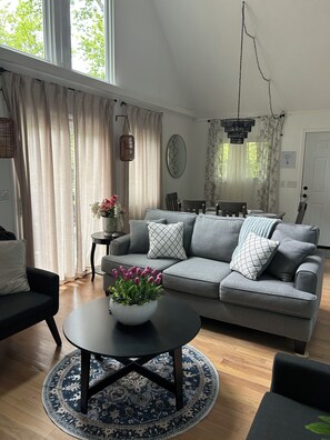 Ein modernes Wohnzimmer mit grauem Sofa, einem runden Couchtisch mit floraler Dekoration, einem Essbereich mit einer Chandelierleuchtre und einem großen Fenster mit Vorhängen.