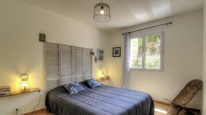 Ein Schlafzimmer mit Bett, einem Fenster mit Vorhängen, einem Sessel und einem Regal mit Büchern und einer Lampe.