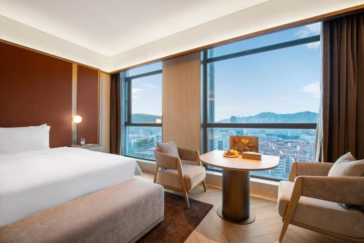 Hotellihuone, jossa on suuri sänky, kaksi nojatuolia, pieni pyöreä pöytä, jossa on hedelmiä, ja näkymä kaupunkiin suurista ikkunoista.