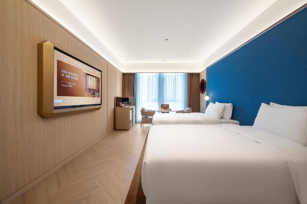 A hotel room with two beds, a TV mounted on the wall, a desk with a chair, and a window with curtains.