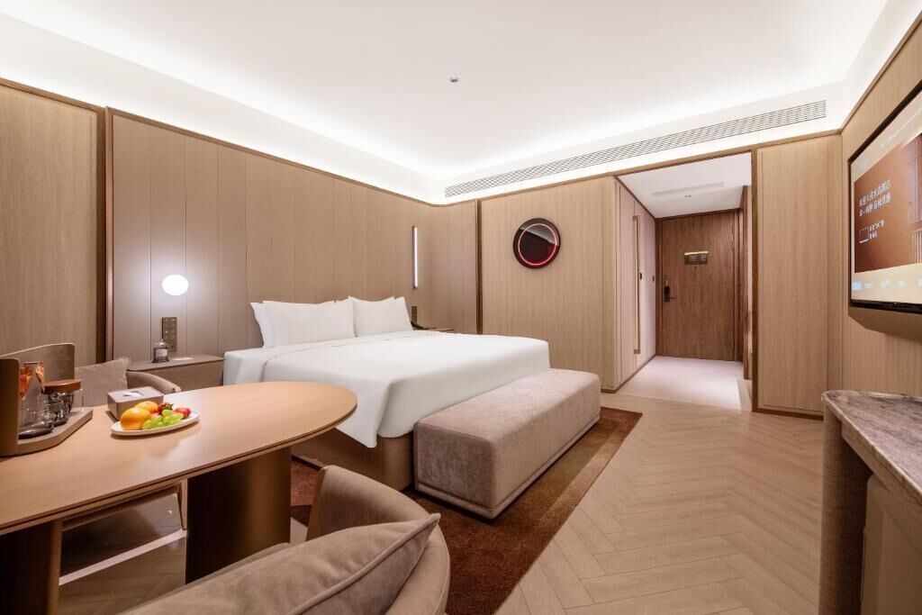 Une chambre d’hôtel moderne avec un lit, une table à manger avec des fruits, une télévision et une salle de bain visible à travers une porte ouverte.