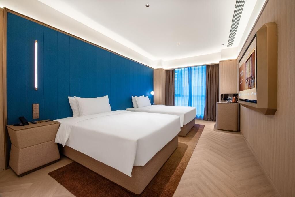 Ein Hotelzimmer mit zwei Betten, einem Nachttisch, einem Fernseher und einem großen Fenster mit Vorhängen.