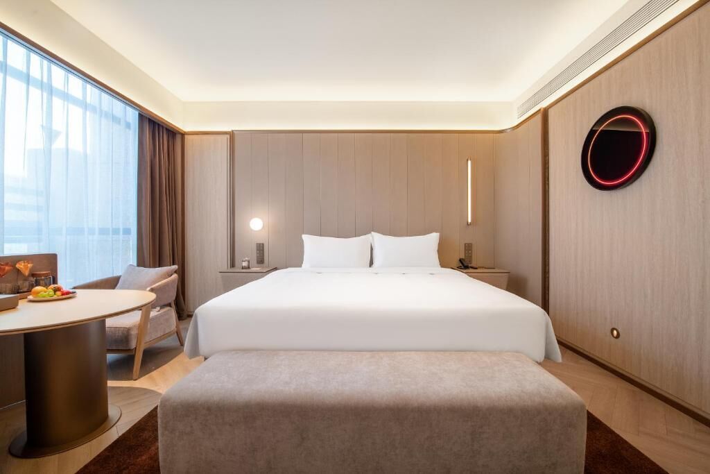 A hotel room with a large bed, bedside tables, a round mirror, a desk with a chair, and a view of the outdoors.