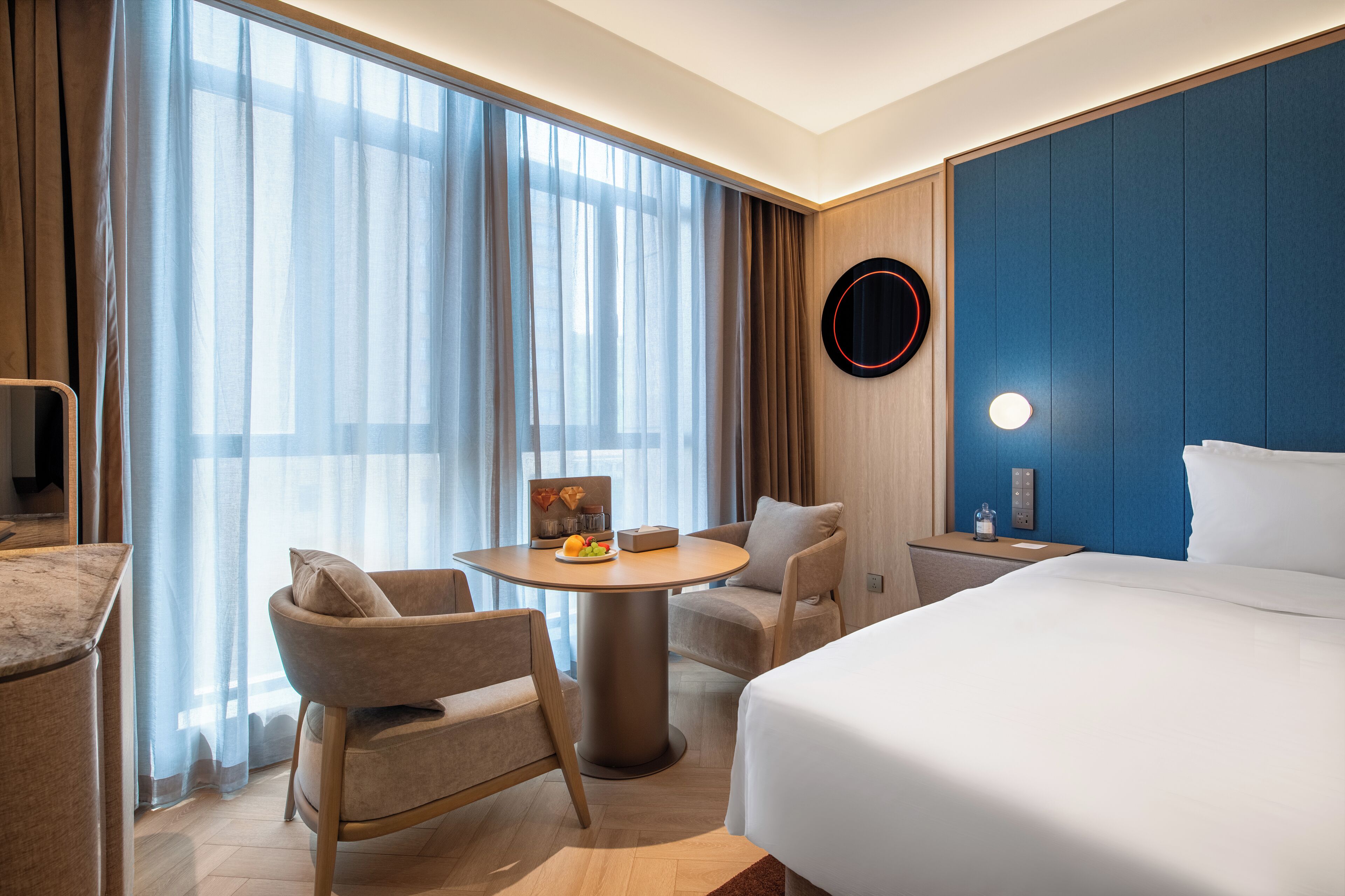 Habitación de hotel con una cama, dos sillones, una mesa redonda con frutero, un espejo circular y una lámpara de pared.