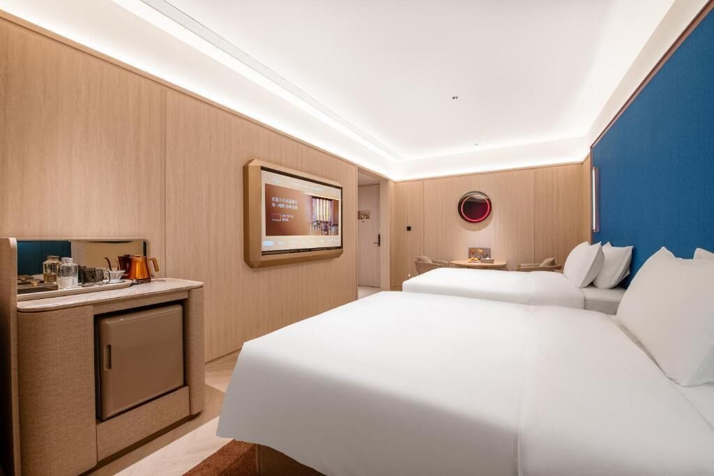 A hotel room with a bed, a desk with a computer, a small kitchenette, and a TV mounted on the wall.