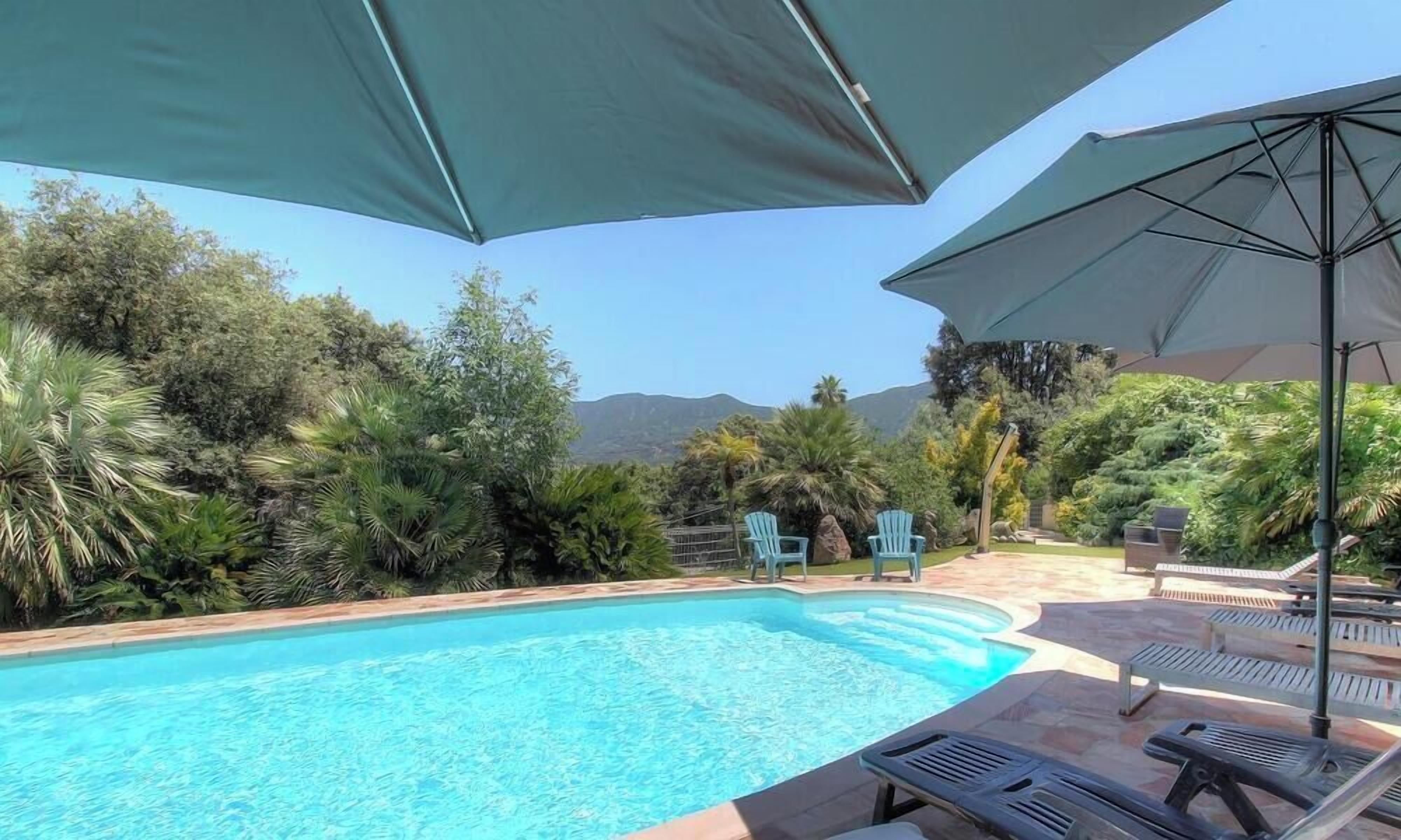 Piscina com espreguiçadeiras e guarda-sóis, cercada por vegetação e montanhas ao fundo.