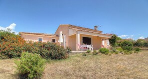 A two-story building with a tiled roof, a covered patio with chairs and a table, and a garden with various plants and flowers. - L'abri côtier - SAGONE - Gîte (SAGONE)