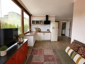 A compact kitchen with a TV, cabinets, a refrigerator, and a window. - Condessa Novo - Storytellers Boutique Villas (São Pedro)