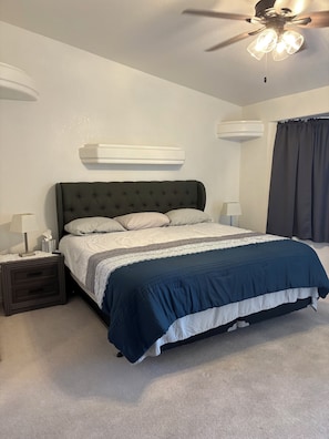 Quarto com cama, mesas de cabeceira, ventilador de teto e janela com cortinas.