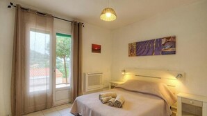Een slaapkamer met een bed, een raam met gordijnen, een radiator, een aan de muur hangend schilderij en een plafondlamp.
