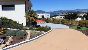 Une maison moderne dotée d’une allée, d’un jardin et offrant une vue sur une ville côtière et des montagnes.